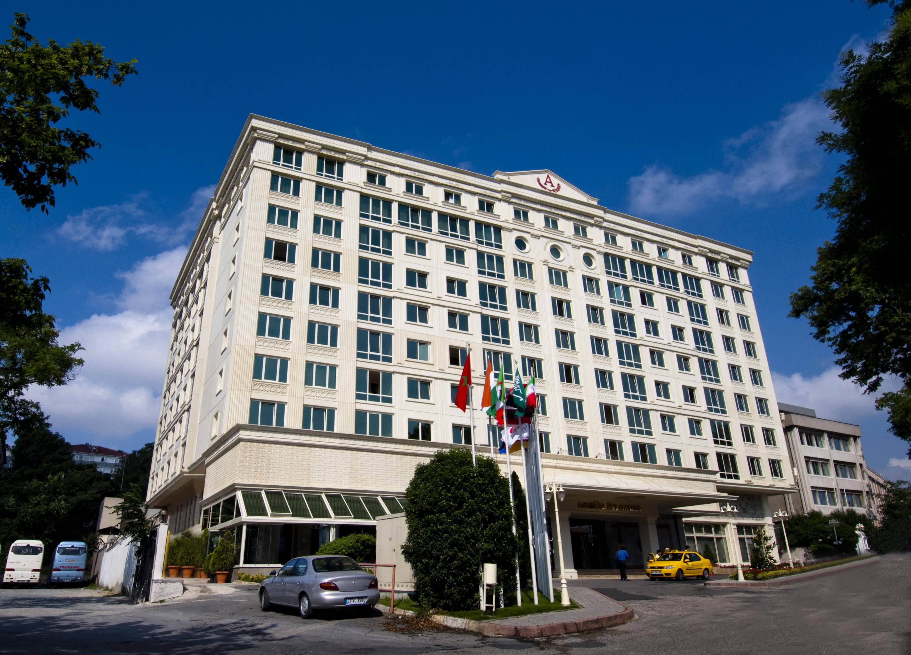 Akgun Istanbul Hotel Exterior foto The image shows a multi-story hotel building with a modern architectural design. The structure has a light-colored façade and large windows. In front of the hotel, there are several flags displayed, and a few cars are parked nearby. The surrounding a