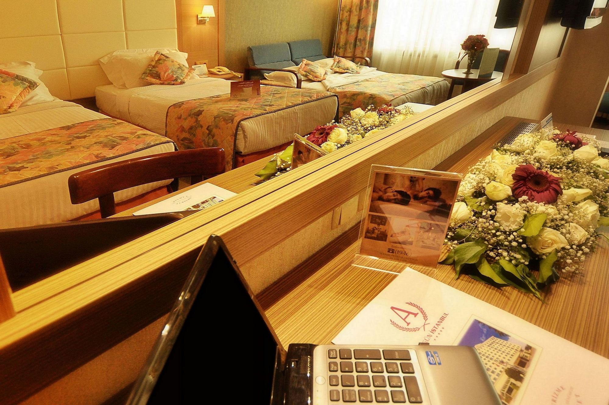 Akgun Istanbul Hotel Exterior foto The photo shows a hotel room with two beds in the background, which are covered with floral bedspreads. In the foreground, there is a wooden table with a laptop placed on it. The table also has a bouquet of flowers and some documents or brochures. A 
