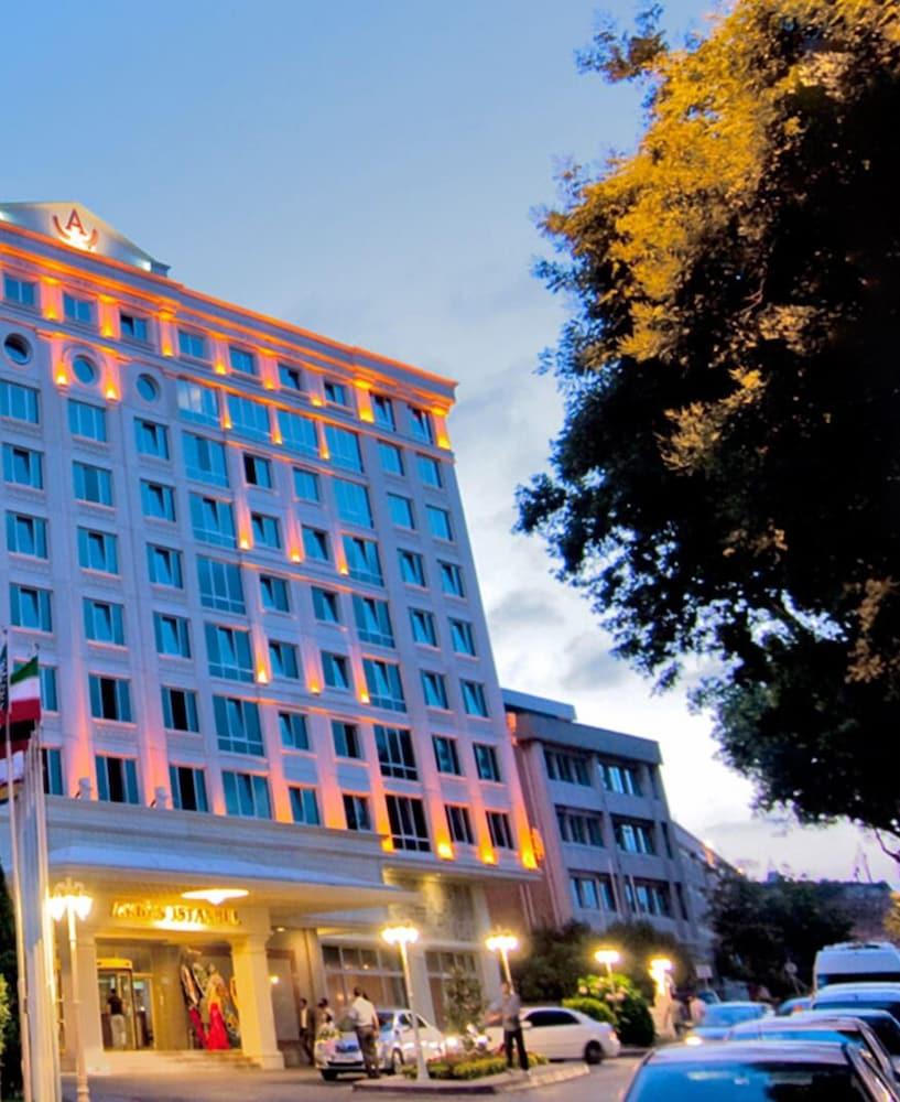 Akgun Istanbul Hotel Exterior foto The photo shows a modern hotel building illuminated with orange lights along its facade. The structure features a series of large windows and a stylized architectural design. In the foreground, there are trees providing some foliage, and flags can be