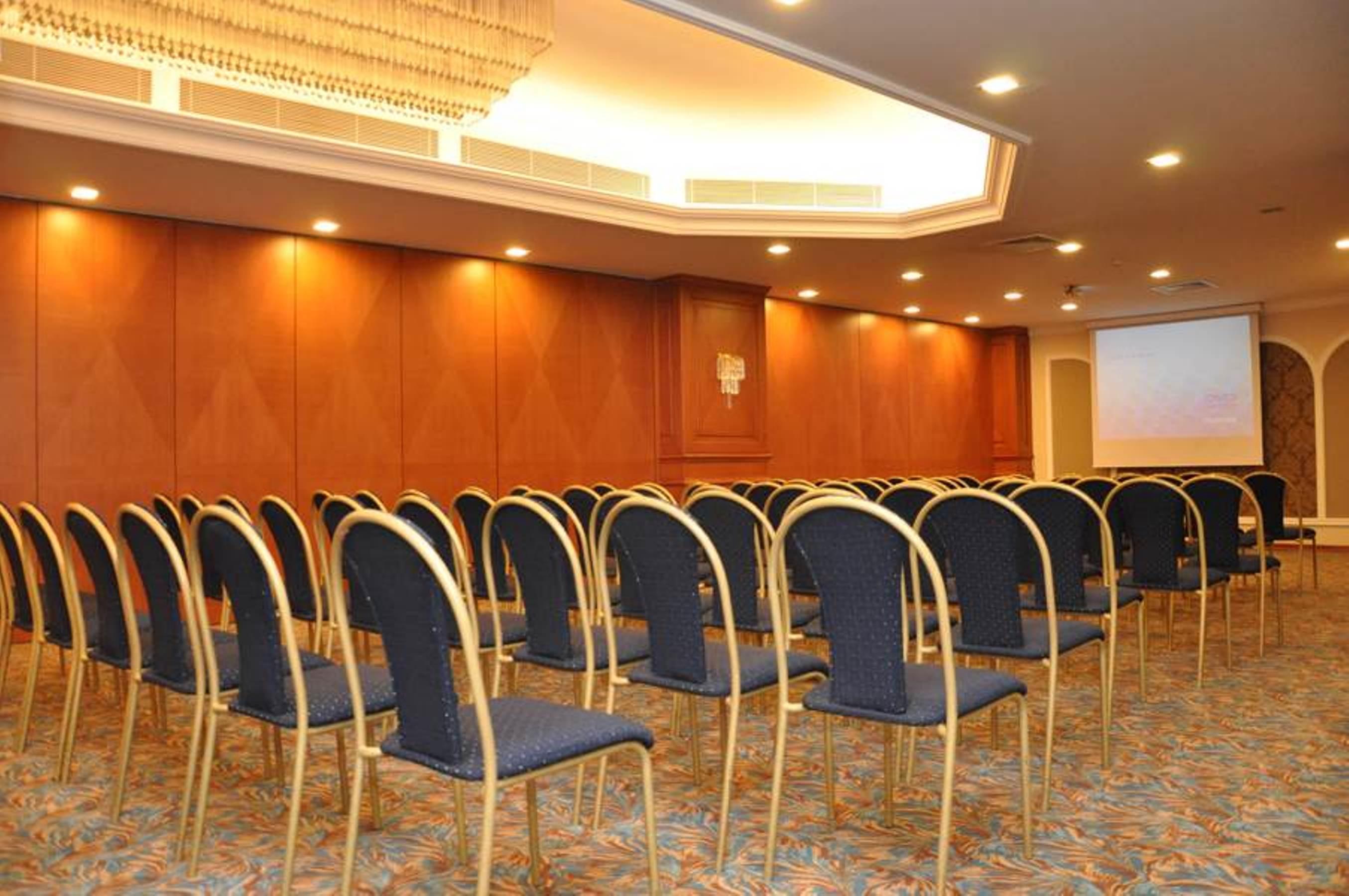 Akgun Istanbul Hotel Exterior foto The photo shows an interior setting, likely a conference or meeting room. There are several rows of blue chairs arranged in a semi-circle facing a screen at the front of the room. The walls are a warm wood color, and there is overhead lighting that c