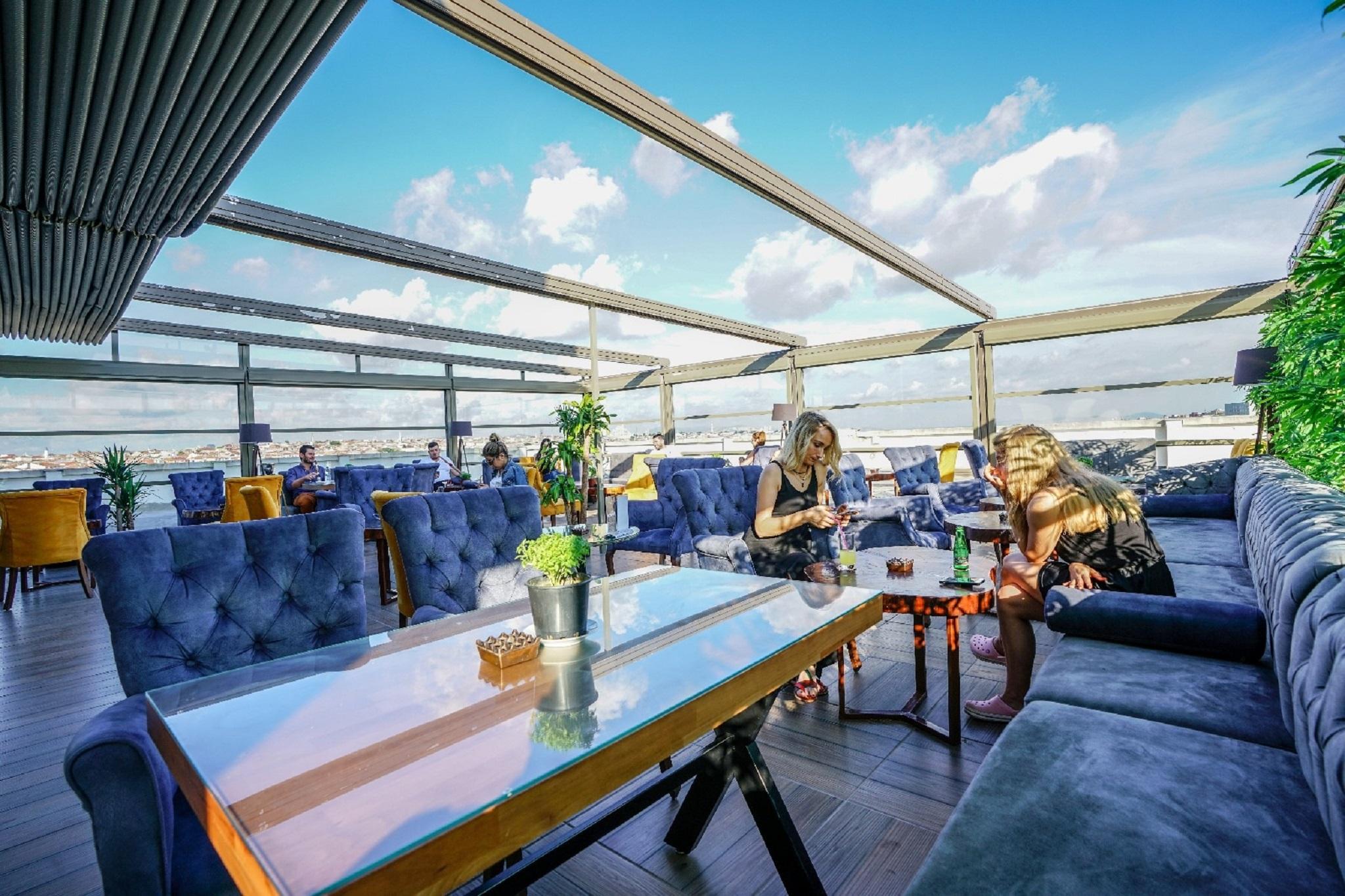 Akgun Istanbul Hotel Exterior foto The photo shows a stylish rooftop lounge or café area with a modern design. There are plush, blue seating arrangements and wooden tables. Two women are engaged in conversation or activities at a table, surrounded by plants and a bright, open atmosphe