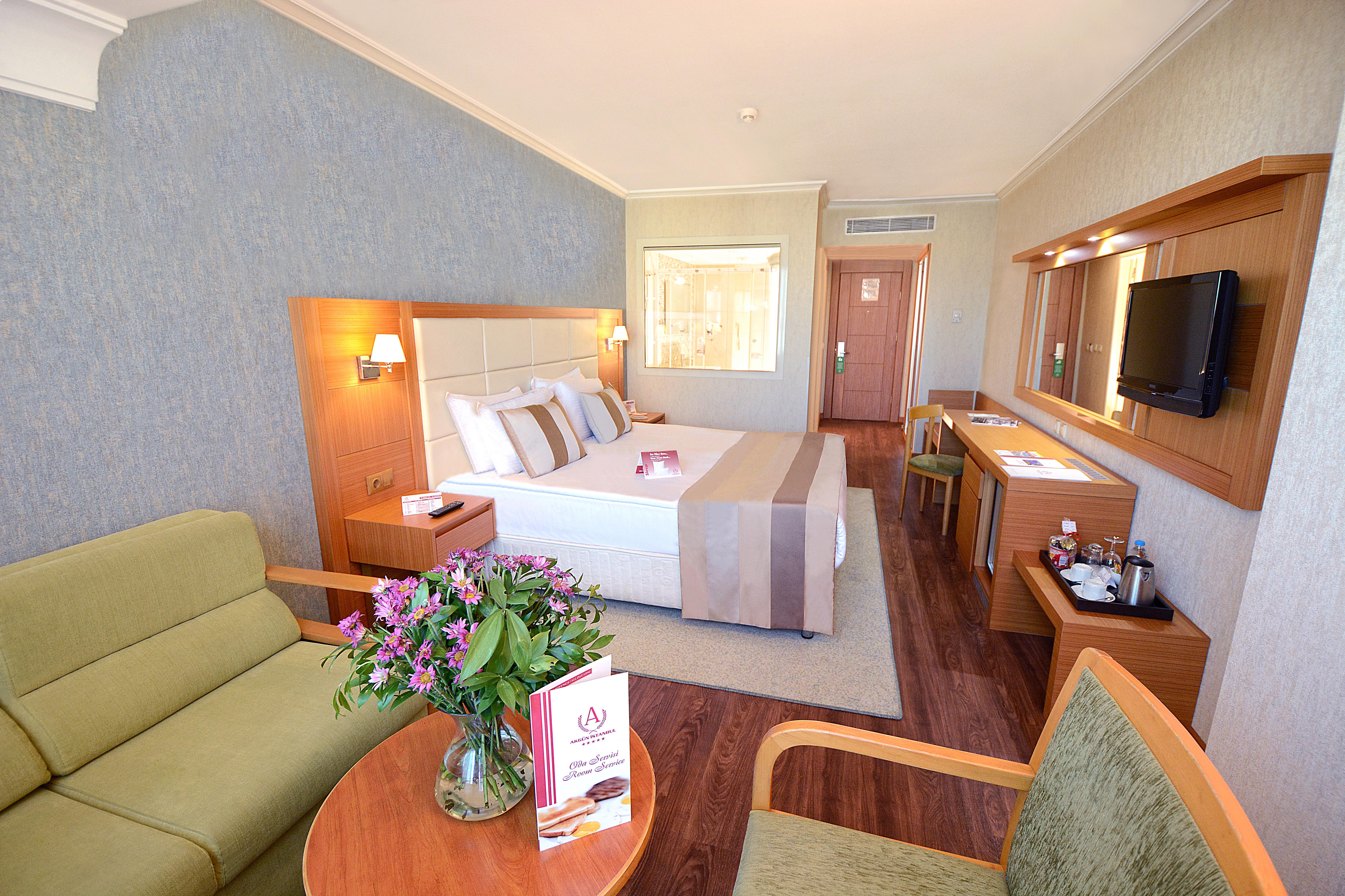 Akgun Istanbul Hotel Exterior foto The image shows a cozy hotel room featuring a spacious double bed with decorative pillows. To the left, there is a small seating area with a green sofa and a wooden chair. A round table holds a vase of flowers and a brochure. On the right side, there