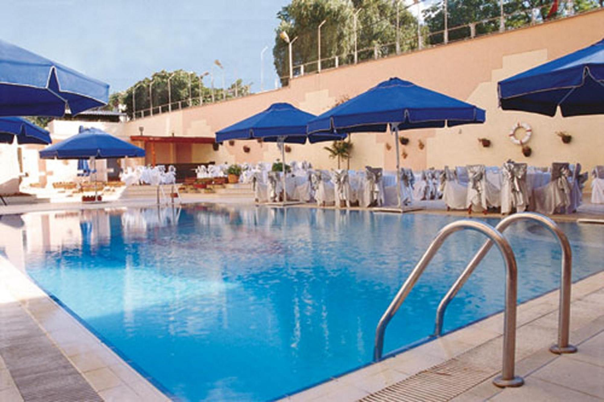 Akgun Istanbul Hotel Exterior foto The photo shows a swimming pool area. The pool is large and filled with clear blue water. Surrounding the pool are several blue umbrellas providing shade. There are also white chairs and tables set up, possibly for events or relaxation. The area is w