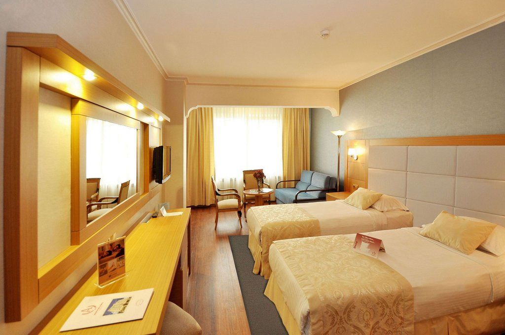 Akgun Istanbul Hotel Exterior foto The photo shows a hotel room featuring two twin beds covered with light-colored bedding. There is a wooden table and a chair near the window, which is draped with light curtains allowing natural light to enter the room. A mirror with built-in lightin